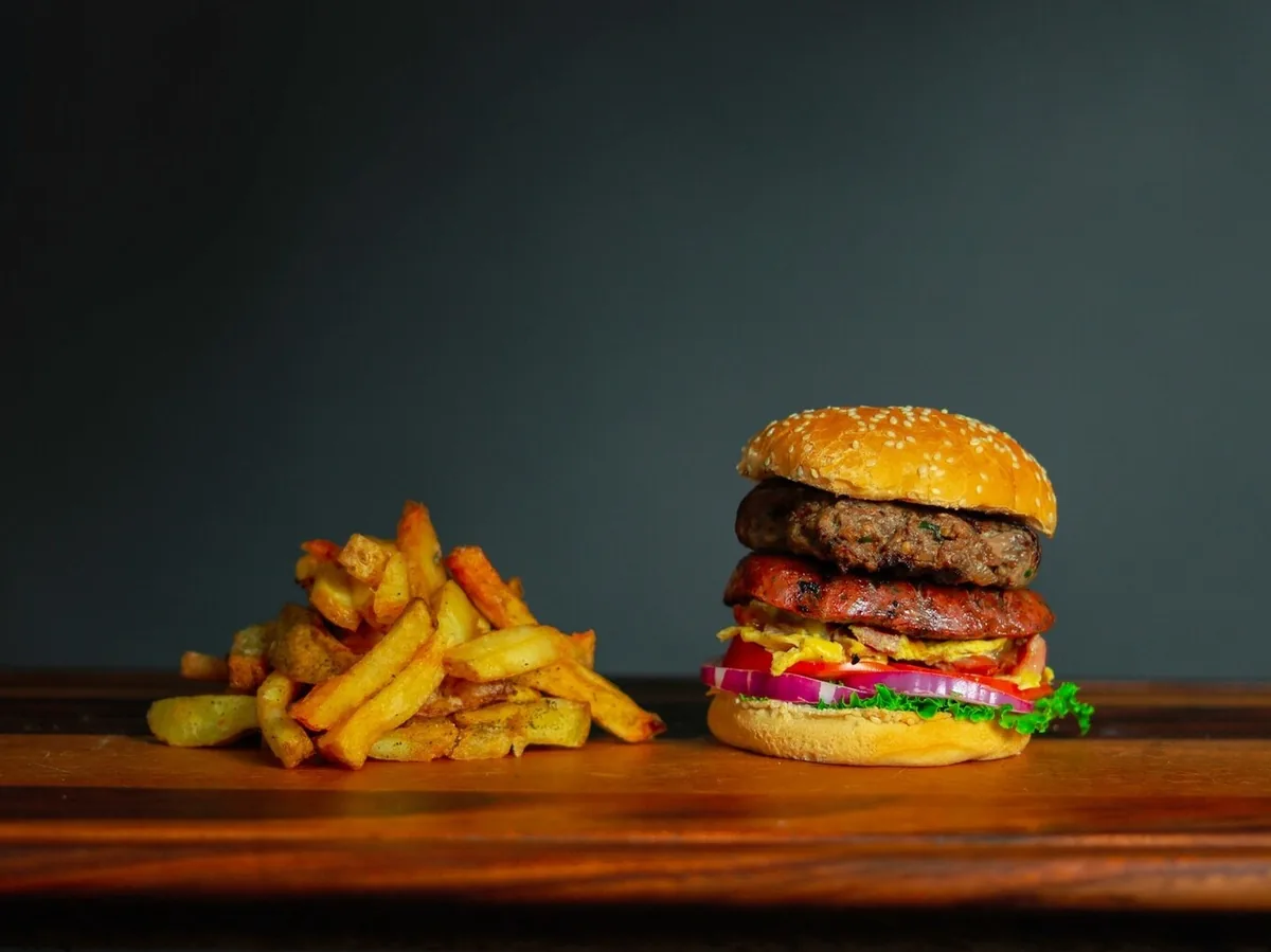A double burger beside a pile of French fries on a wooden table against a dark background.