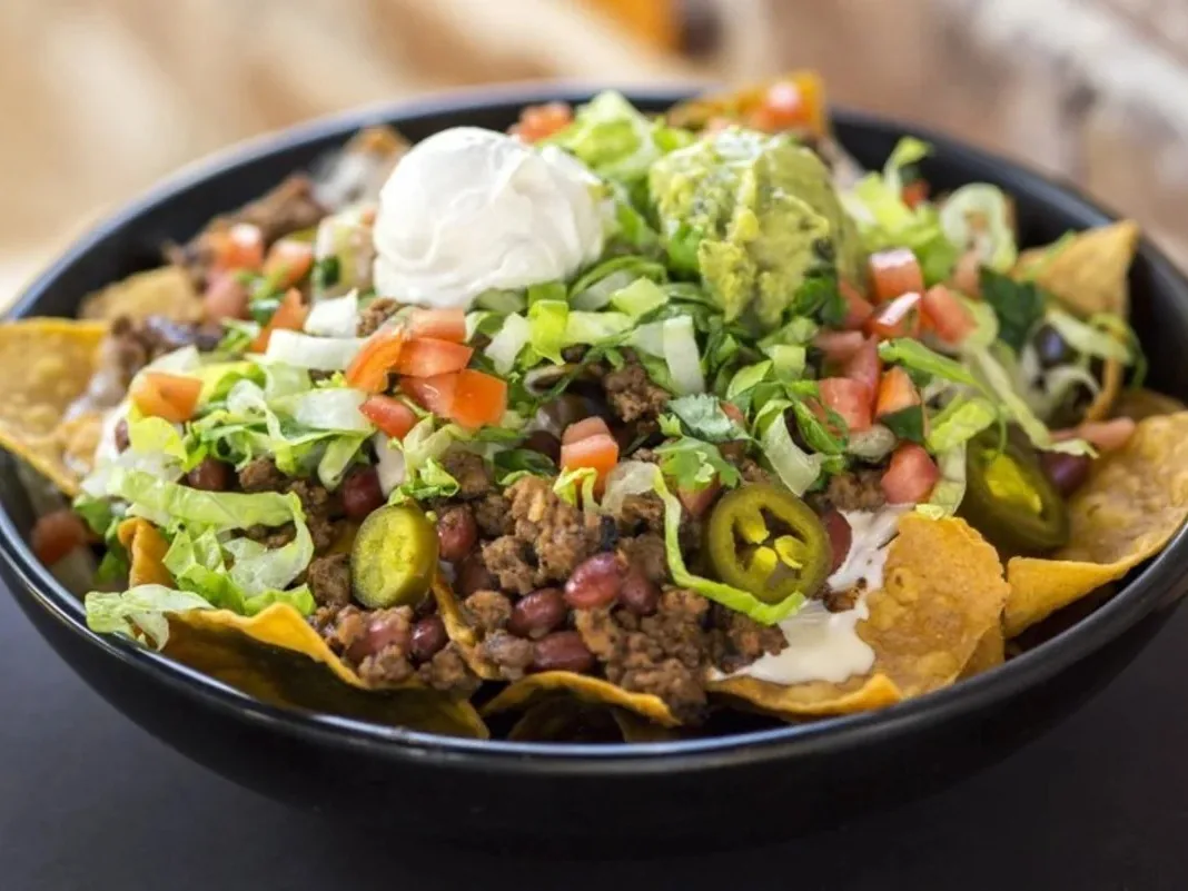 A bowl of nachos topped with ground meat, beans, lettuce, tomatoes, jalapenos, guacamole, and sour cream.