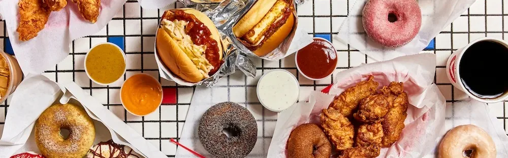 Hot dogs, fried chicken, donuts, dipping sauces and a cup of coffee on a checkered table.