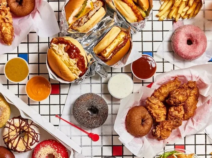 An overhead spread of burgers, fried chicken, donuts, french fries and dipping sauces on a tiled table.