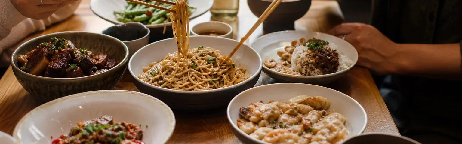Shared bowls of noodles, rice and meat dishes on a table, with chopsticks lifting noodles.