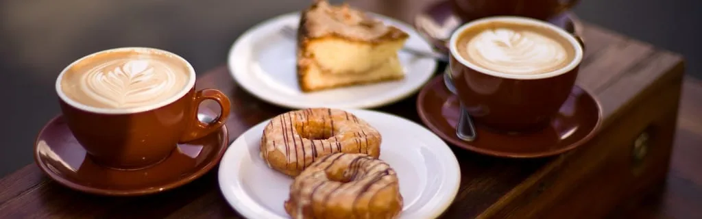 Two cups of coffee with latte art, glazed donuts and a slice of cake on a wooden table.