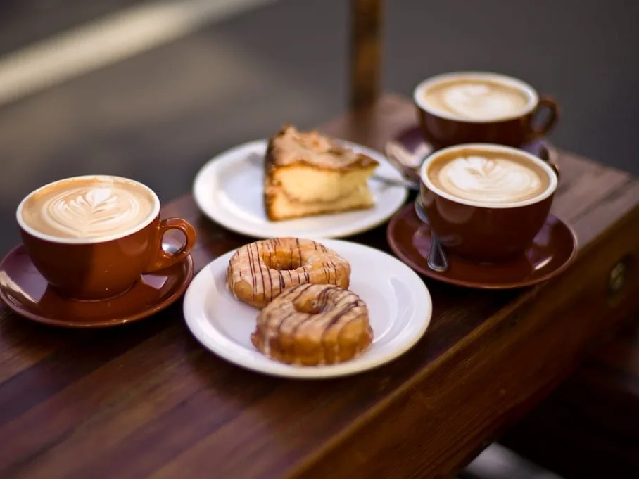 Three cups of coffee with latte art, a slice of cake, and doughnuts on a wooden table.