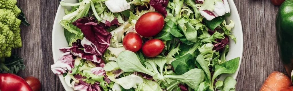 Bowl of mixed salad leaves and cherry tomatoes surrounded by broccoli, carrots and peppers.