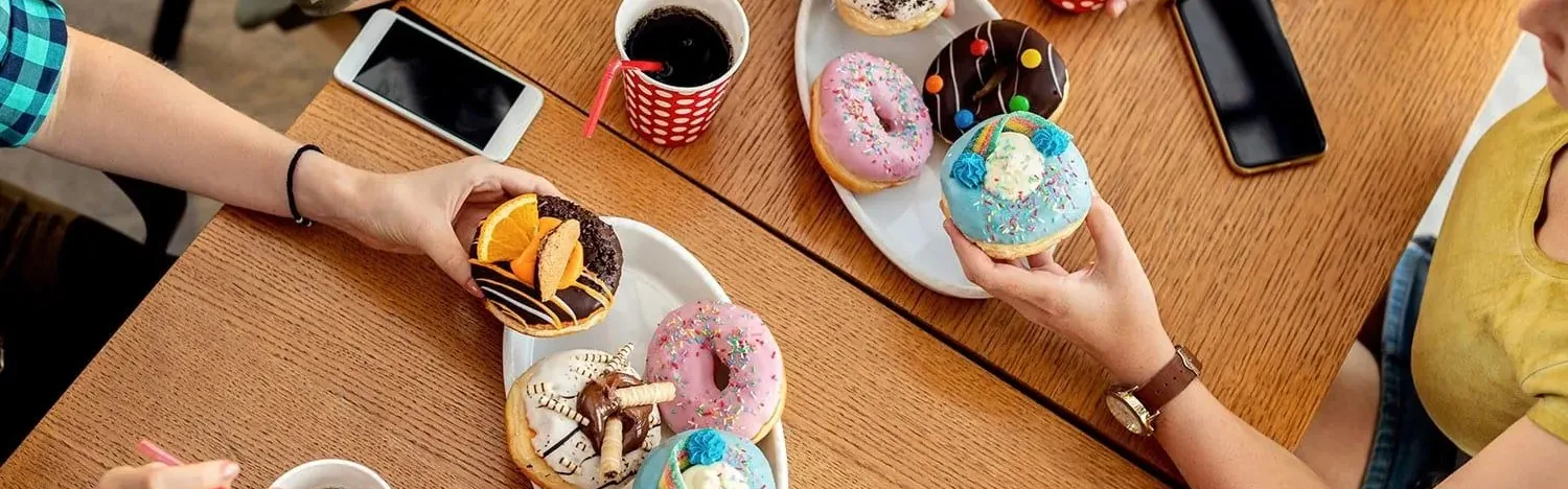 Hands holding colorful frosted donuts over a wooden table with phones and drinks.