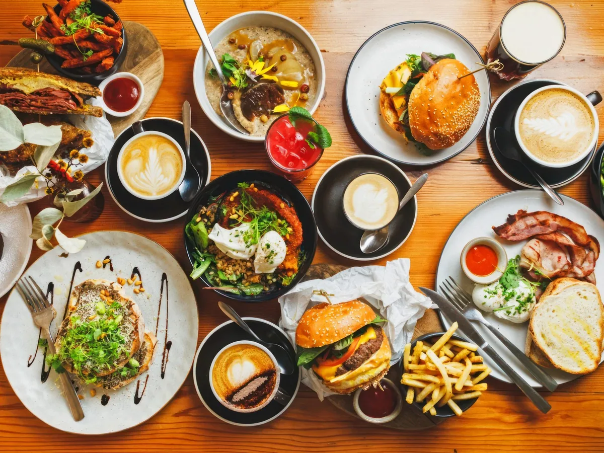An overhead view of a wooden table covered with burgers, French fries, salads, soups, coffee, and drinks.