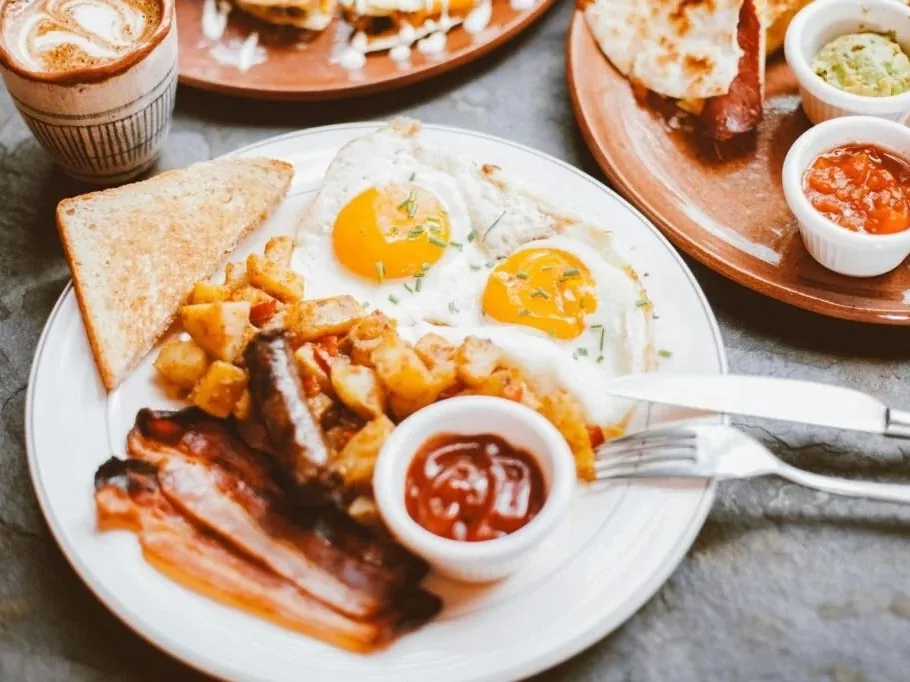 A breakfast plate with fried eggs, bacon, sausage, potatoes and toast beside a cup of coffee.