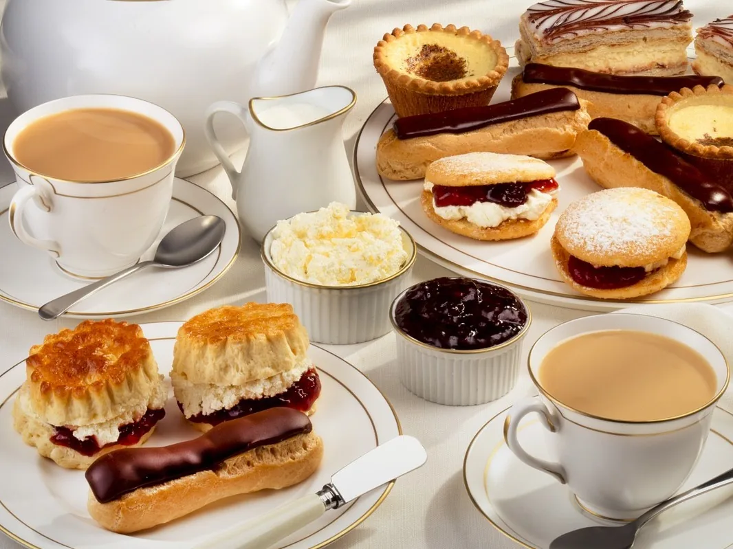 Cups of tea with scones, jam, cream, eclairs, and cakes arranged on plates.