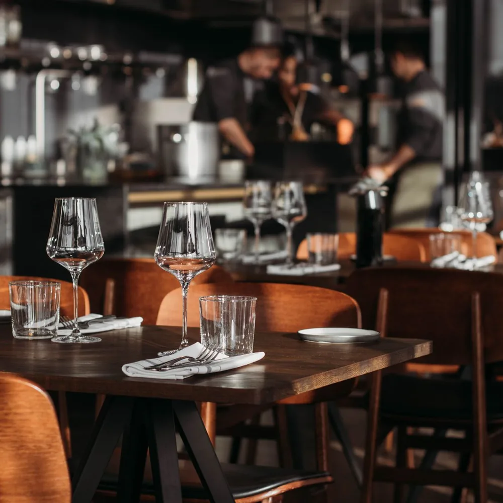 A restaurant dining room with set tables in the foreground and staff working in an open kitchen.