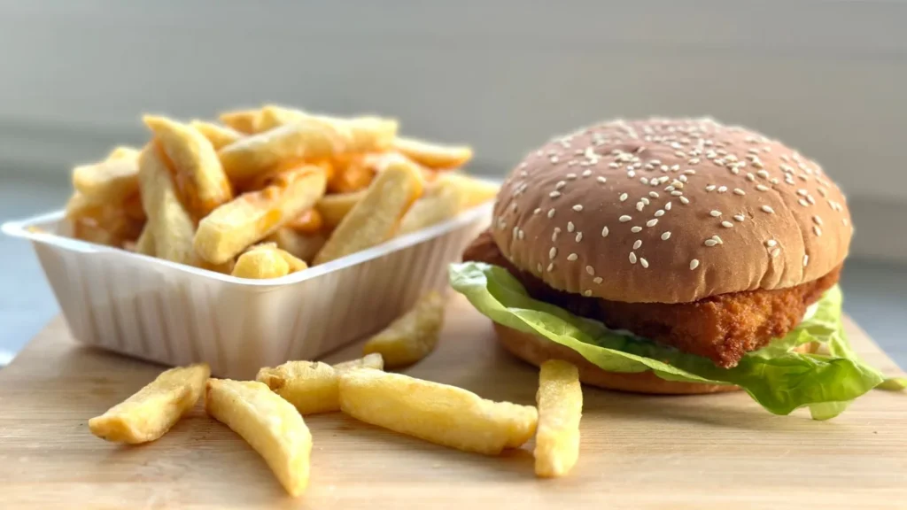 A fried chicken burger with lettuce on a sesame seed bun beside a container of French fries on a wooden board.