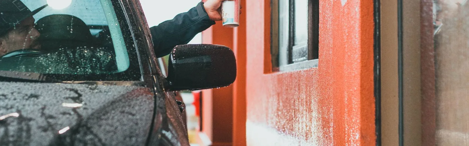 A driver handing a drink cup through a car window at a drive-thru window.