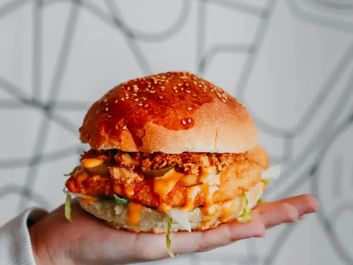 A hand holding a fried chicken sandwich with sauce, pickles, and lettuce on a sesame seed bun.