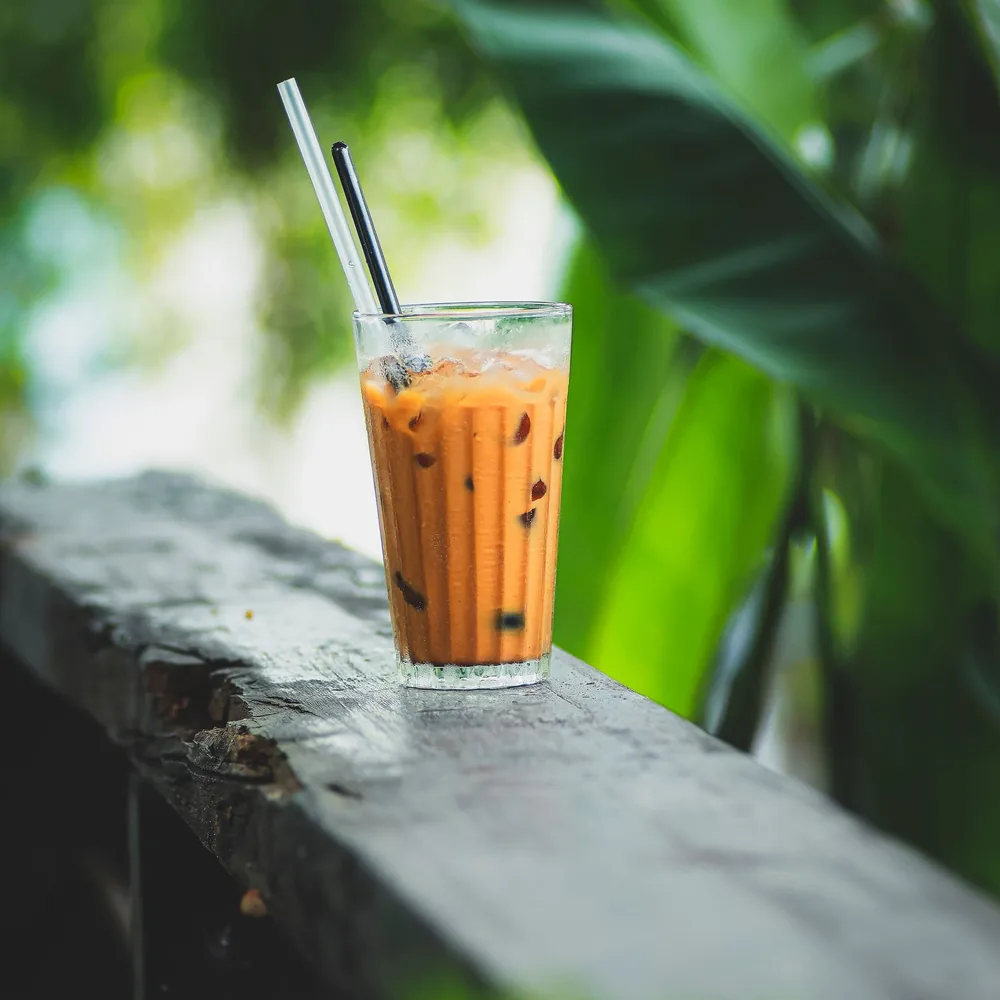 A glass of iced milky tea with straws on a wooden ledge against green leaves.