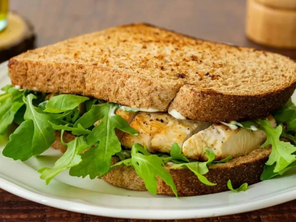 A grilled chicken sandwich on toasted brown bread with fresh arugula on a white plate.