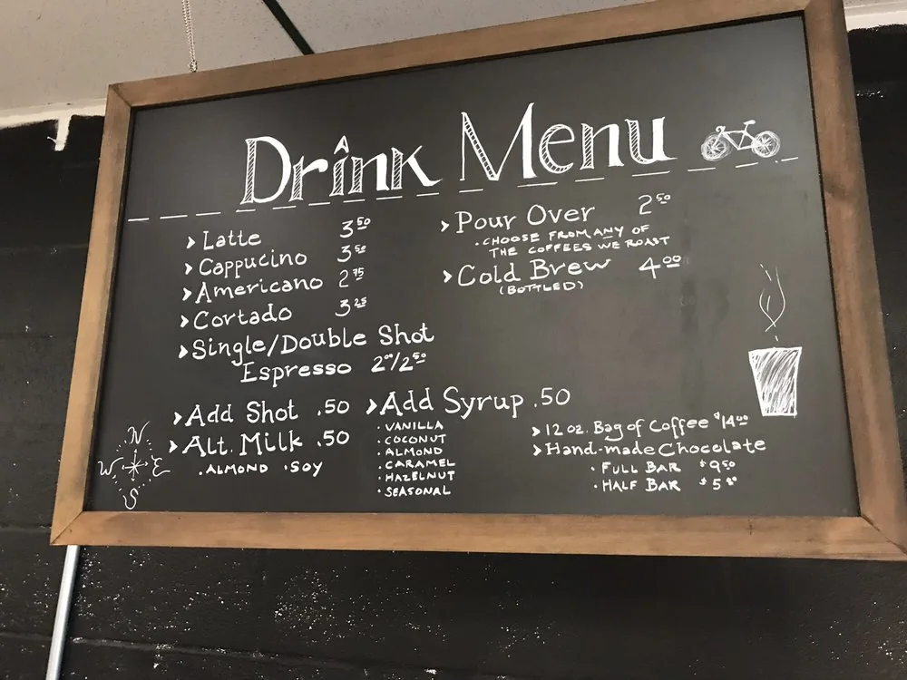A framed chalkboard drink menu with handwritten coffee drinks and prices.
