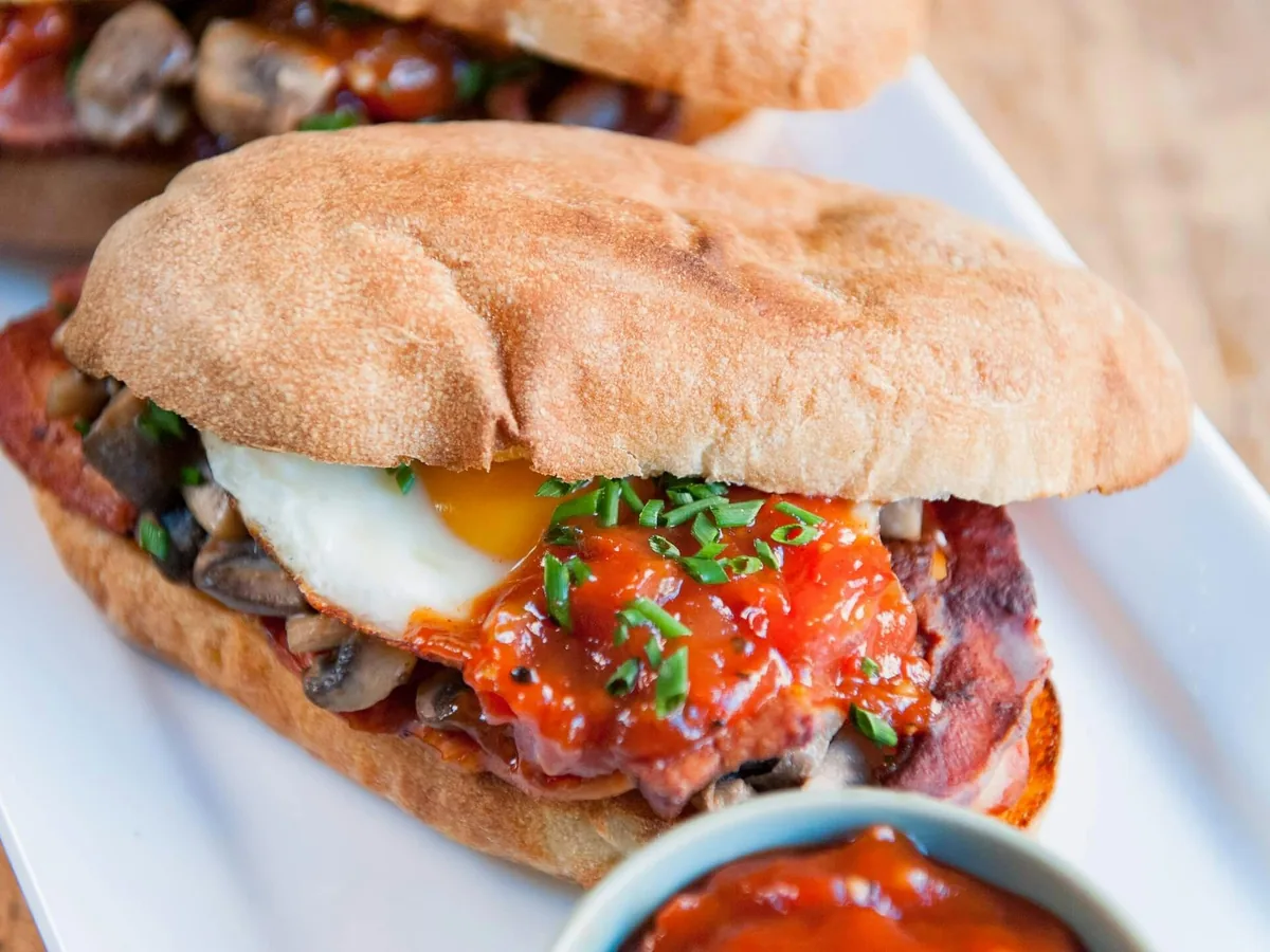 A breakfast roll filled with a fried egg, bacon, mushrooms, and tomato sauce, with a small bowl of sauce beside it.