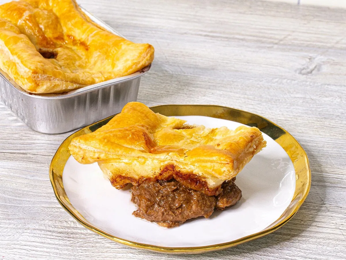 A steak bake with golden pastry and meat filling on a plate, with another bake in a foil tray behind it.