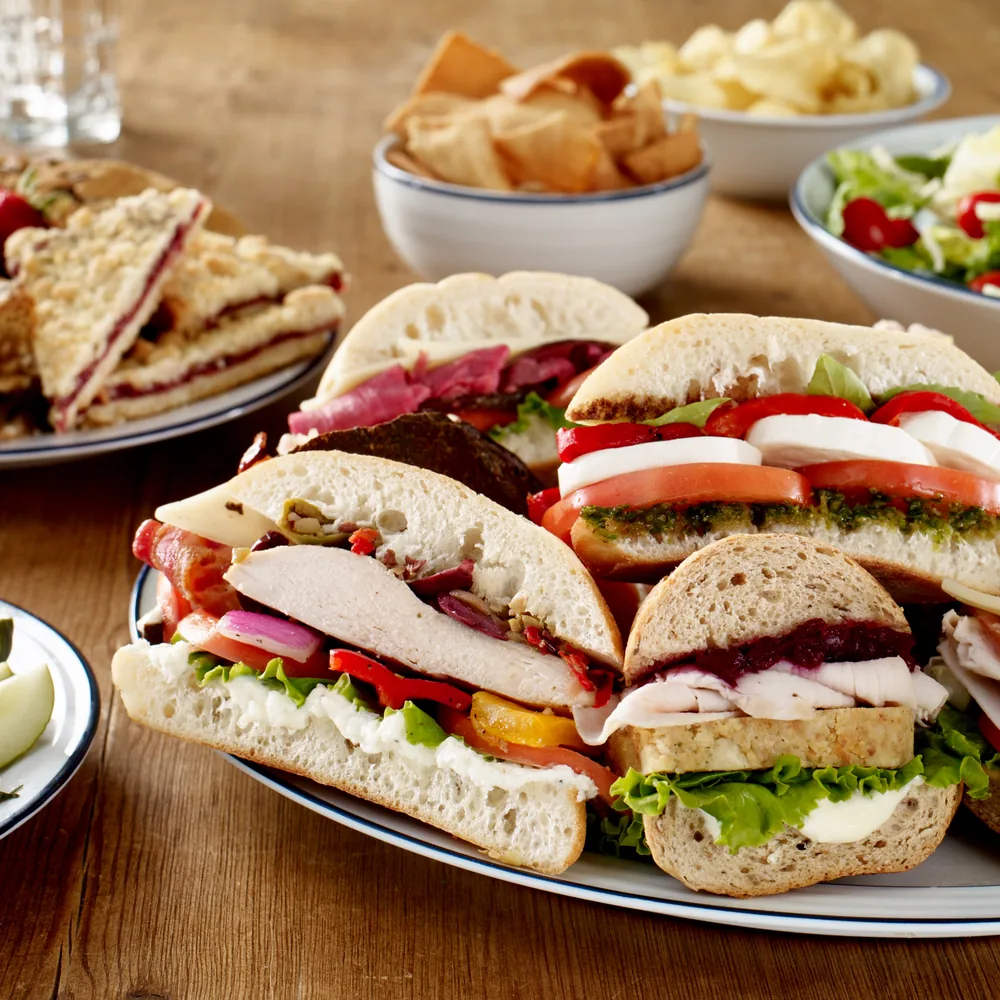 Filled sandwiches with tomato, cheese, and salad on plates, with bowls of crisps and salad behind them.