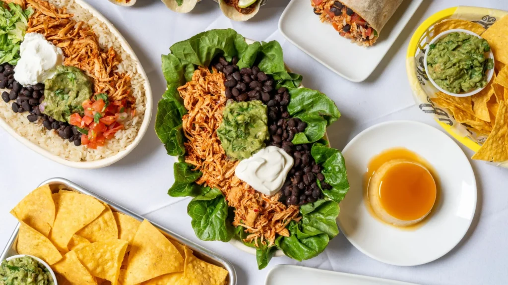 An overhead spread of salads with shredded chicken, beans, lettuce, and guacamole, with tortilla chips, a burrito, and a caramel dessert.