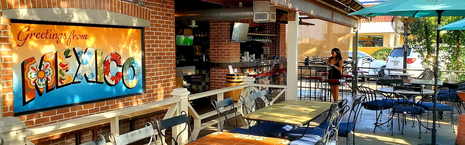 An outdoor restaurant patio with tables, chairs, umbrellas, and a colorful mural on a brick wall.