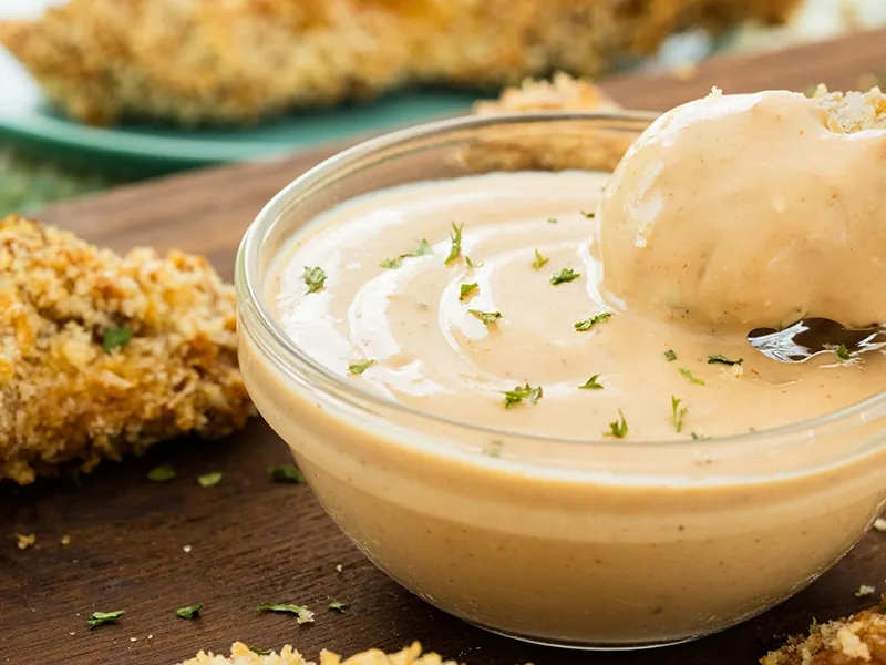 A breaded chicken tender dipped in a bowl of creamy sauce with herbs, with more tenders behind it.