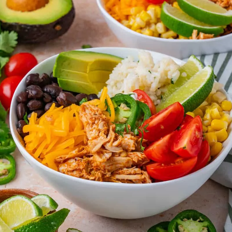 A bowl with shredded chicken, rice, black beans, cheese, tomatoes, avocado, and lime wedges.