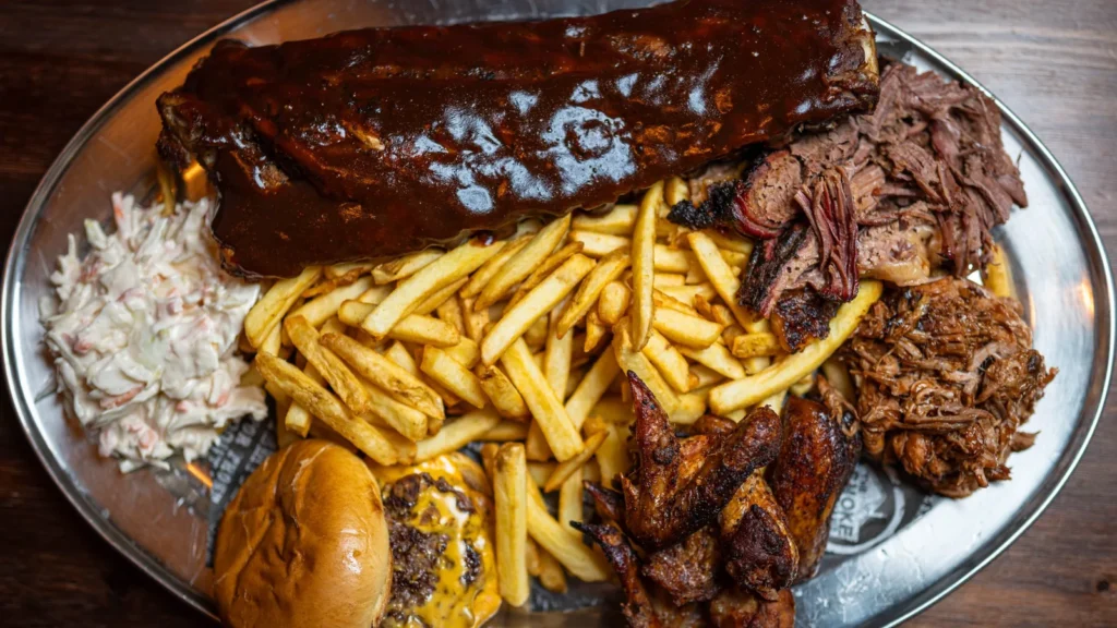 A metal platter with glazed barbecue ribs, pulled meat, French fries, coleslaw, chicken wings, and a cheeseburger.