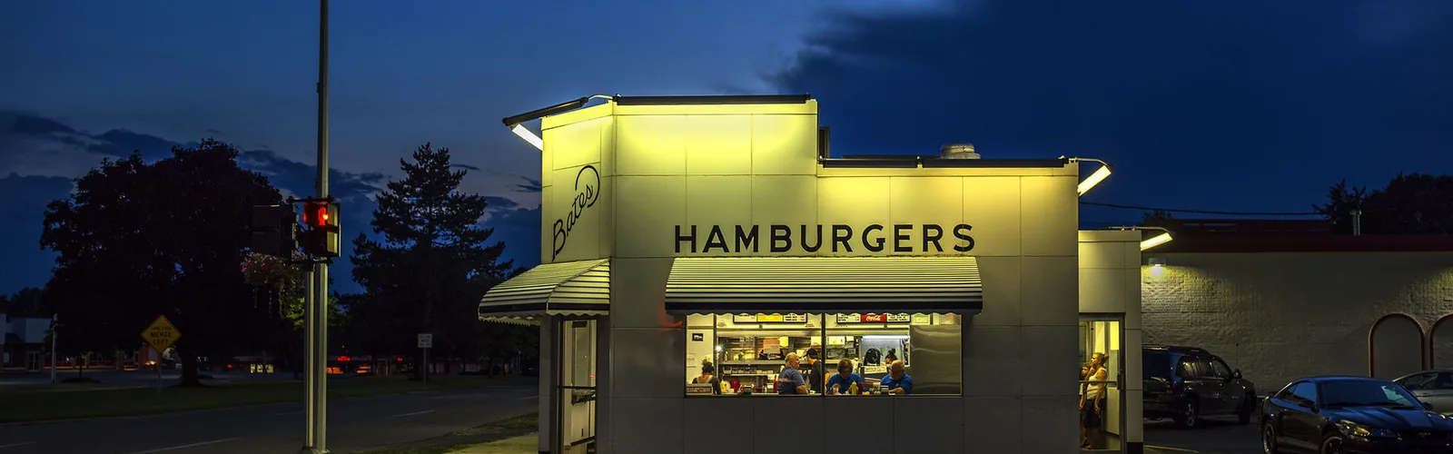 An illuminated hamburger restaurant building at dusk with cars parked outside.