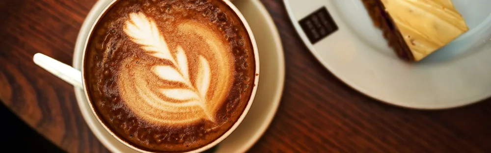 A cup of coffee with leaf-shaped latte art beside a slice of cake on a wooden table.