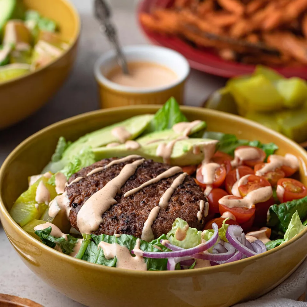 A salad bowl topped with a grilled patty, avocado, cherry tomatoes, red onion, and a drizzle of sauce.