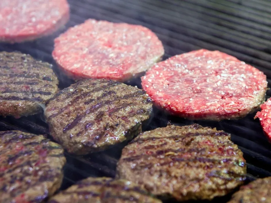 Beef patties with grill marks cooking on a grill, some still pink and others charred.