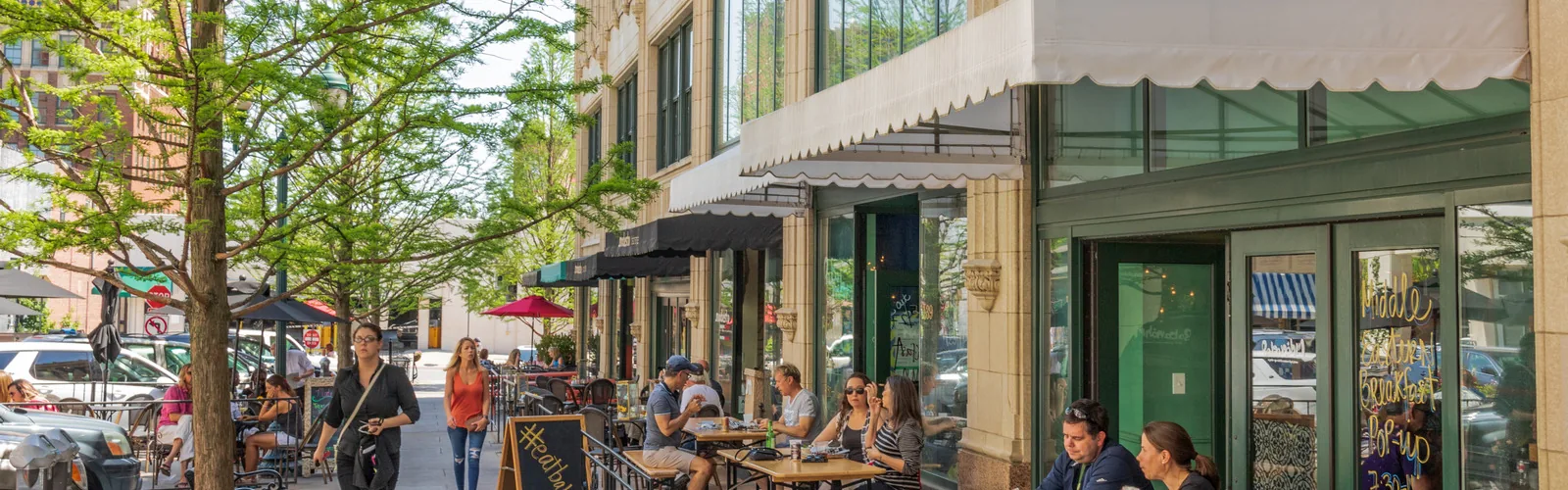 People sitting at outdoor tables in front of a restaurant with large windows on a street.