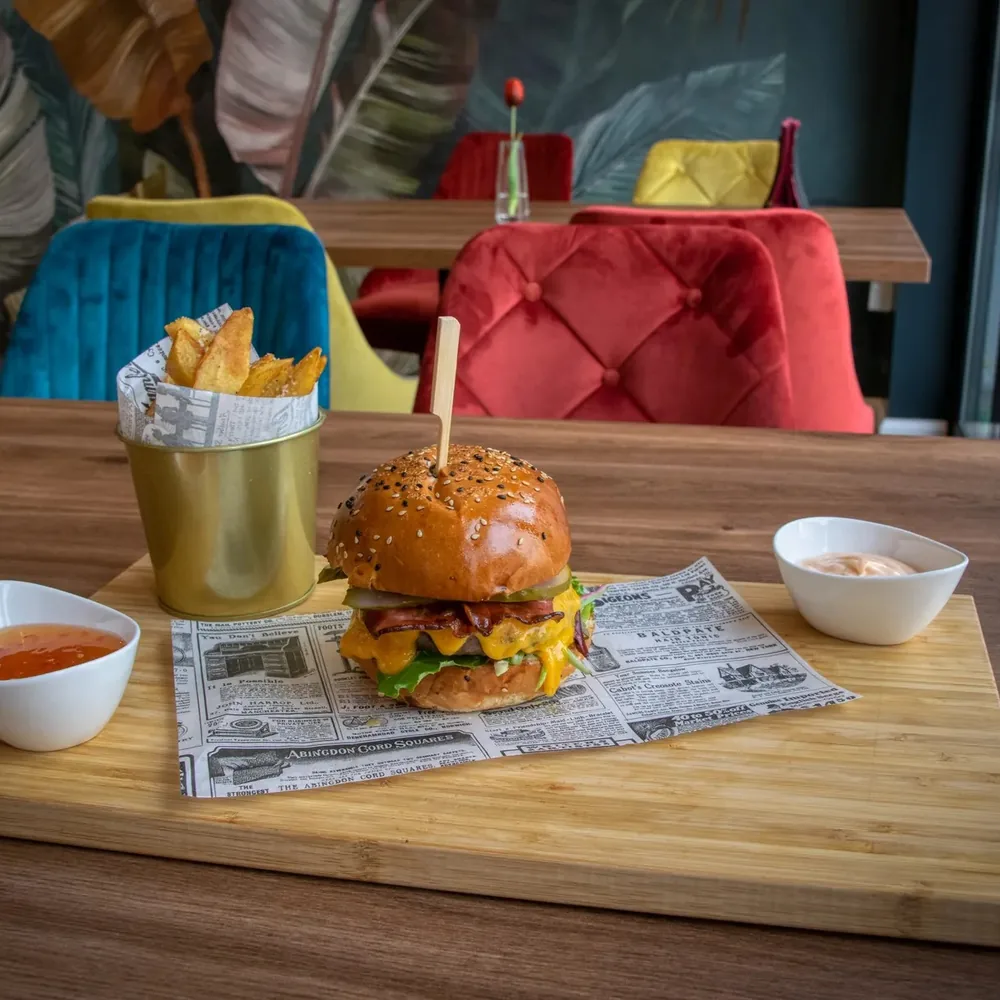 A burger and fries with dipping sauces on a wooden board in a restaurant with colourful chairs.