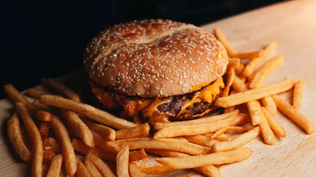 A cheeseburger in a sesame seed bun surrounded by fries on a wooden board.