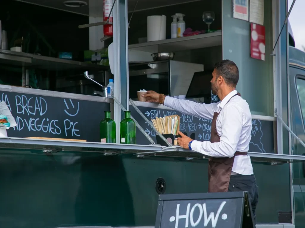 A server handing a cup across the counter of a food truck with a chalkboard menu.