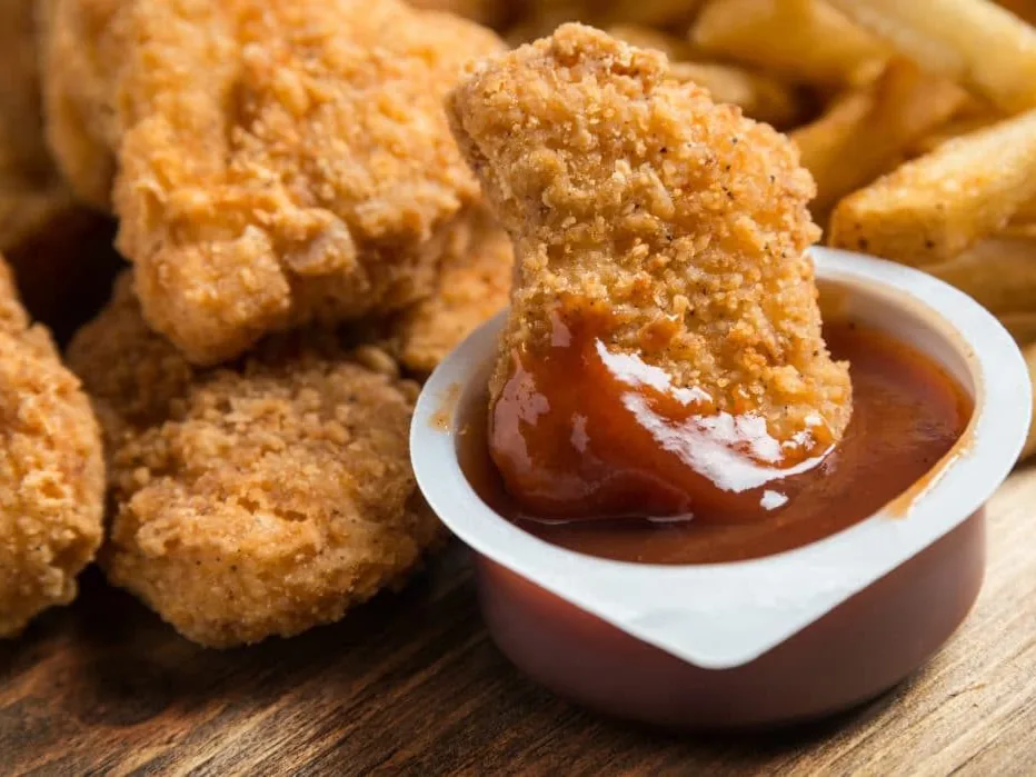 Breaded chicken nuggets with one nugget dipped in sauce and fries in the background.
