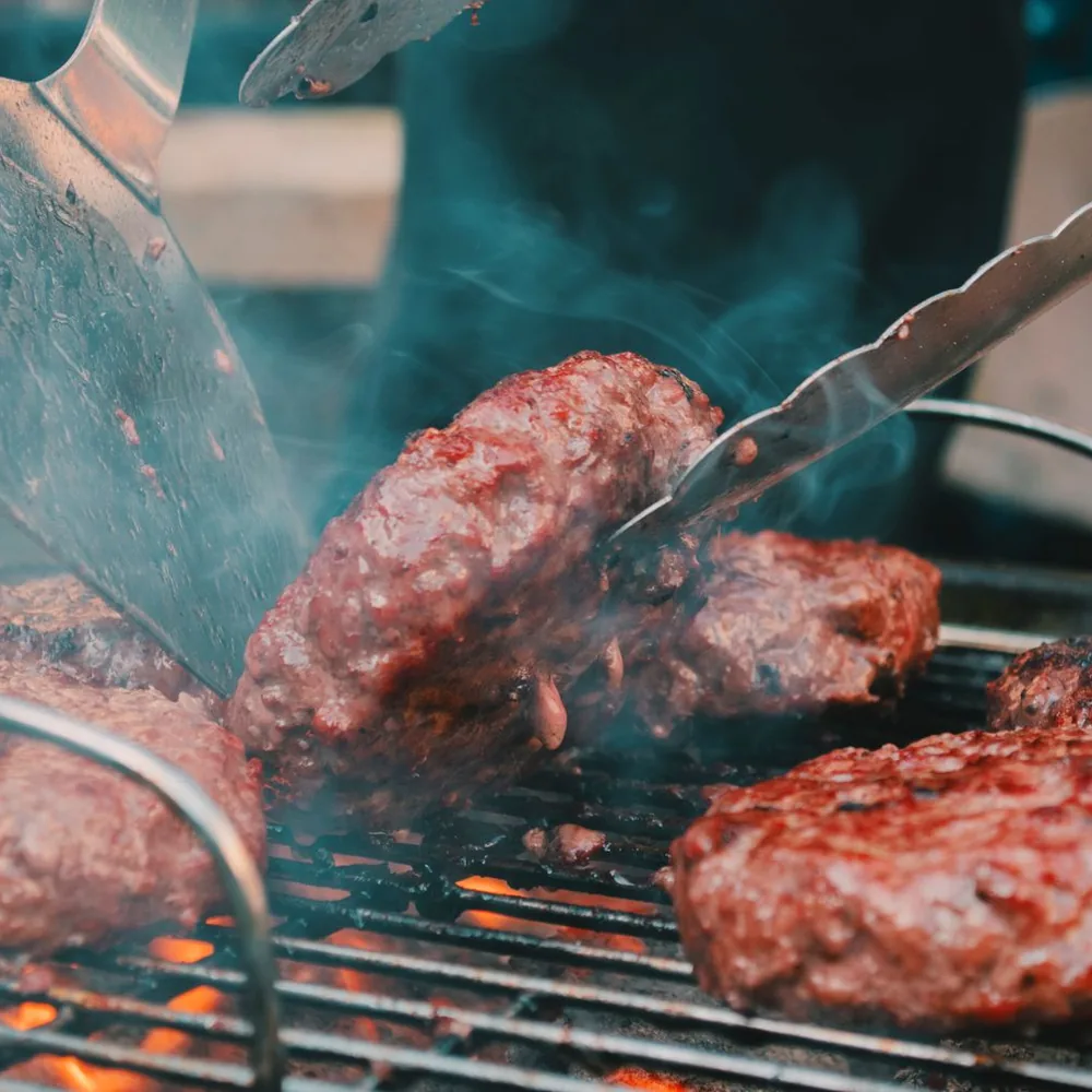 Beef burger patties sizzling on a grill while tongs turn one patty.