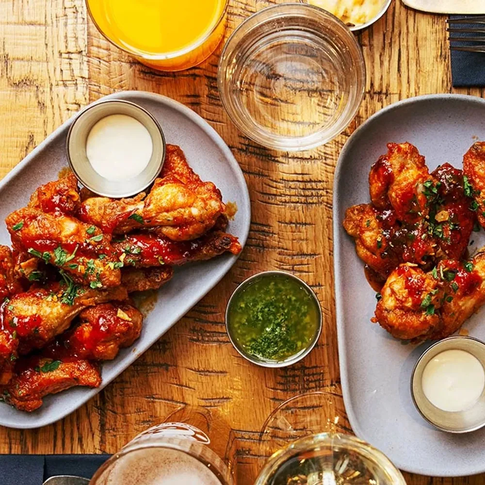 Two plates of glazed chicken wings with small bowls of dipping sauce on a wooden table.