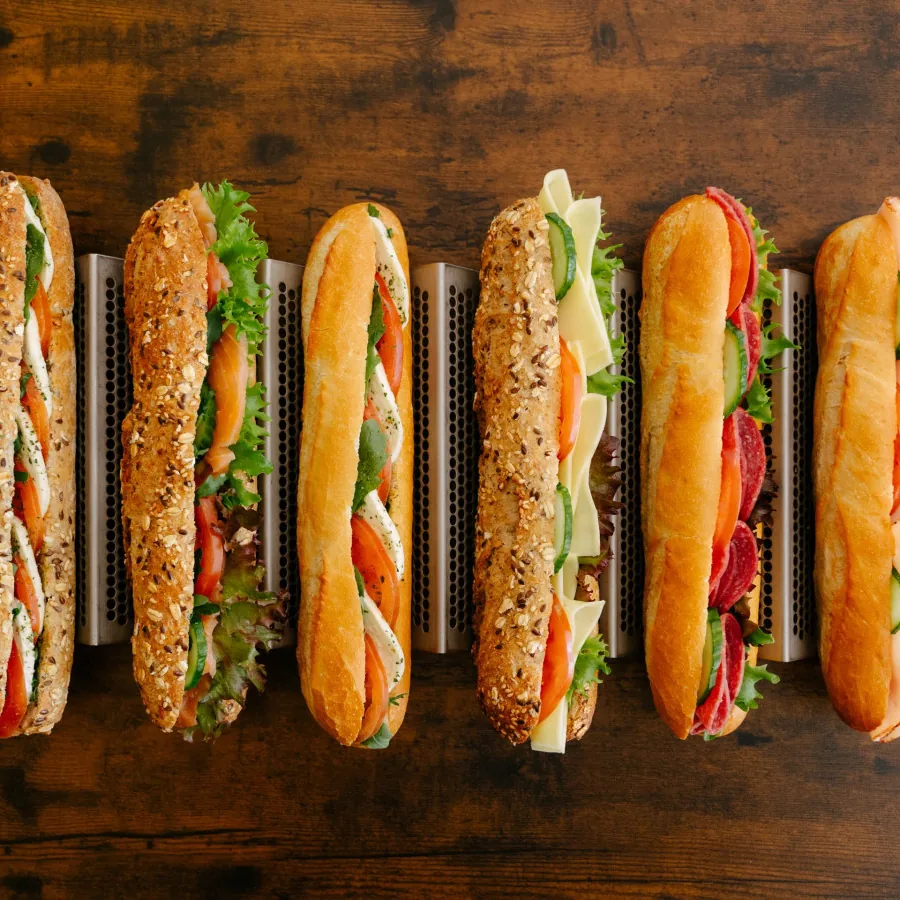 A row of filled sandwiches and baguettes with salad and cheese arranged on a wooden board.