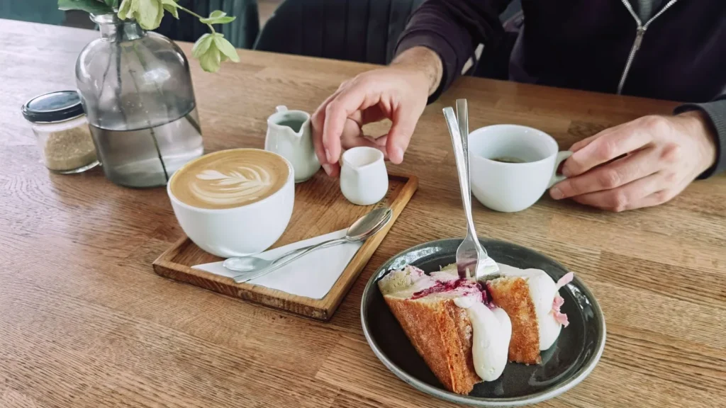 A latte with heart-shaped latte art and a slice of cake with cream served on a wooden café table.