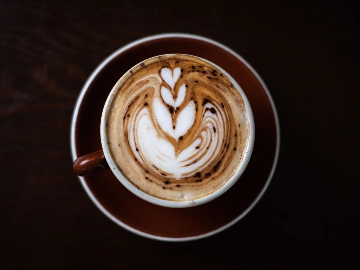 A cup of coffee with leaf-shaped latte art viewed from above on a dark table.