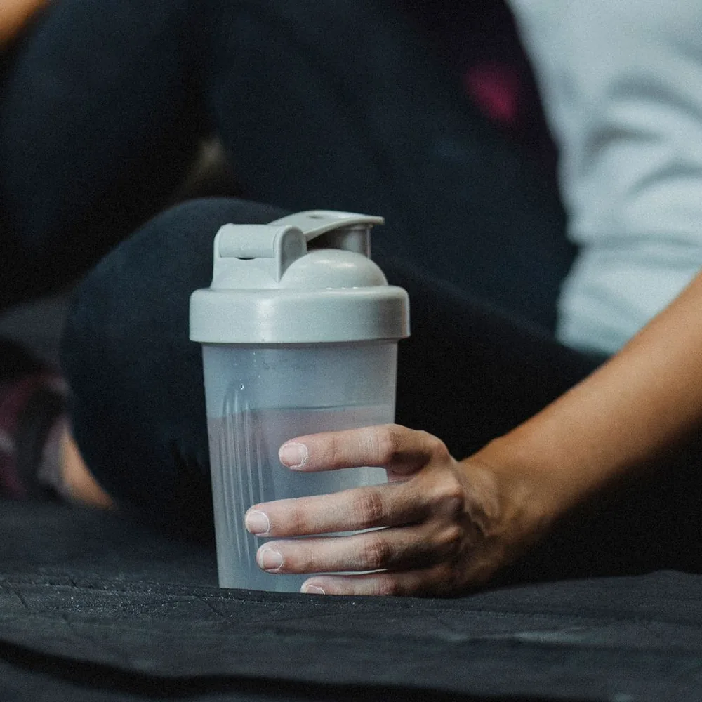 A hand holding a clear shaker bottle beside a person sitting on the floor.