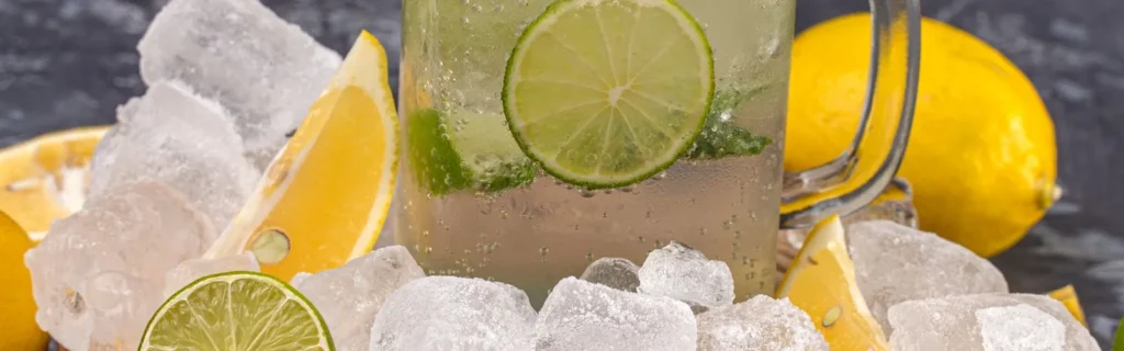 A glass of iced citrus drink with lime slices surrounded by ice, lemons and limes.
