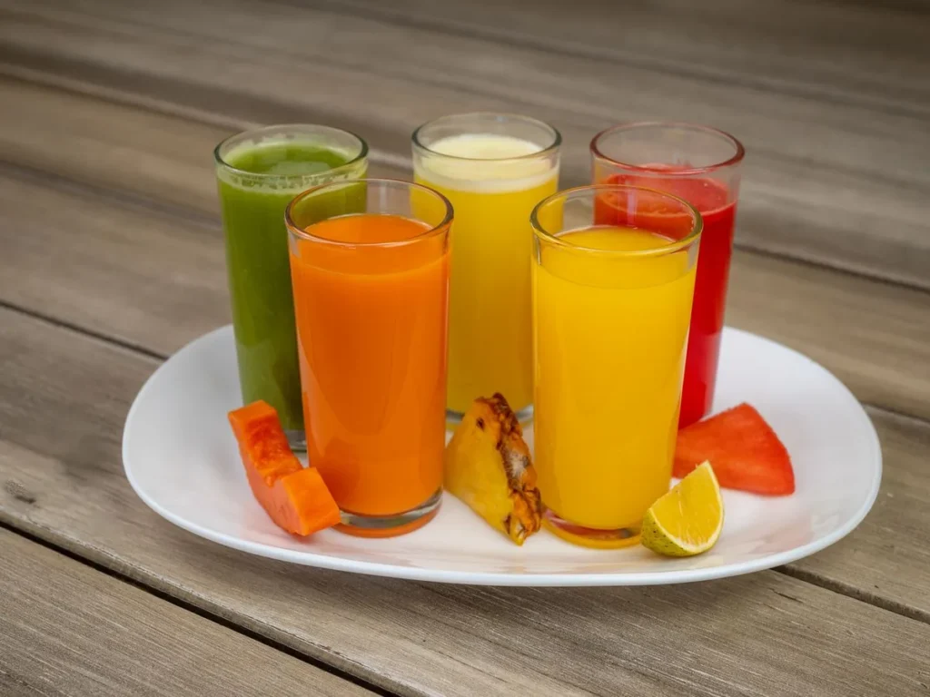 Five glasses of colourful fruit juices with fresh fruit on a plate.