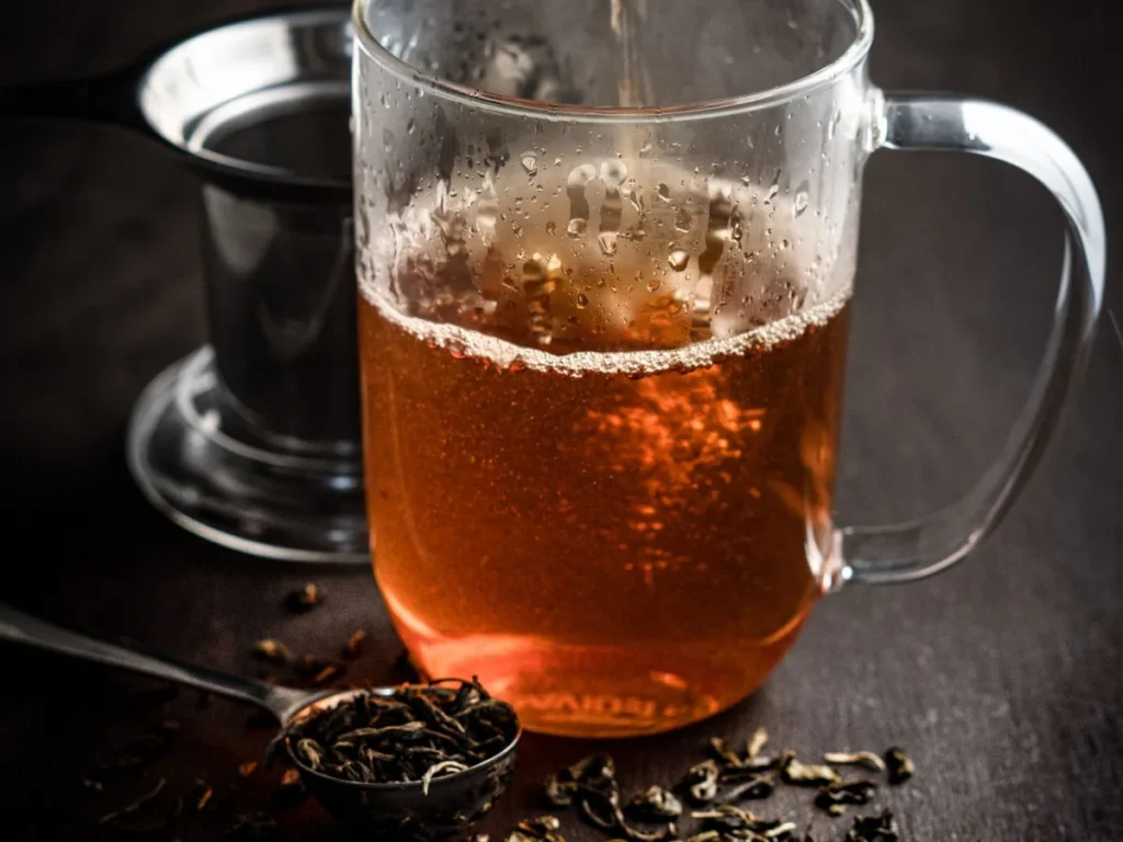 Amber tea being poured into a glass mug beside a spoon of loose tea leaves.