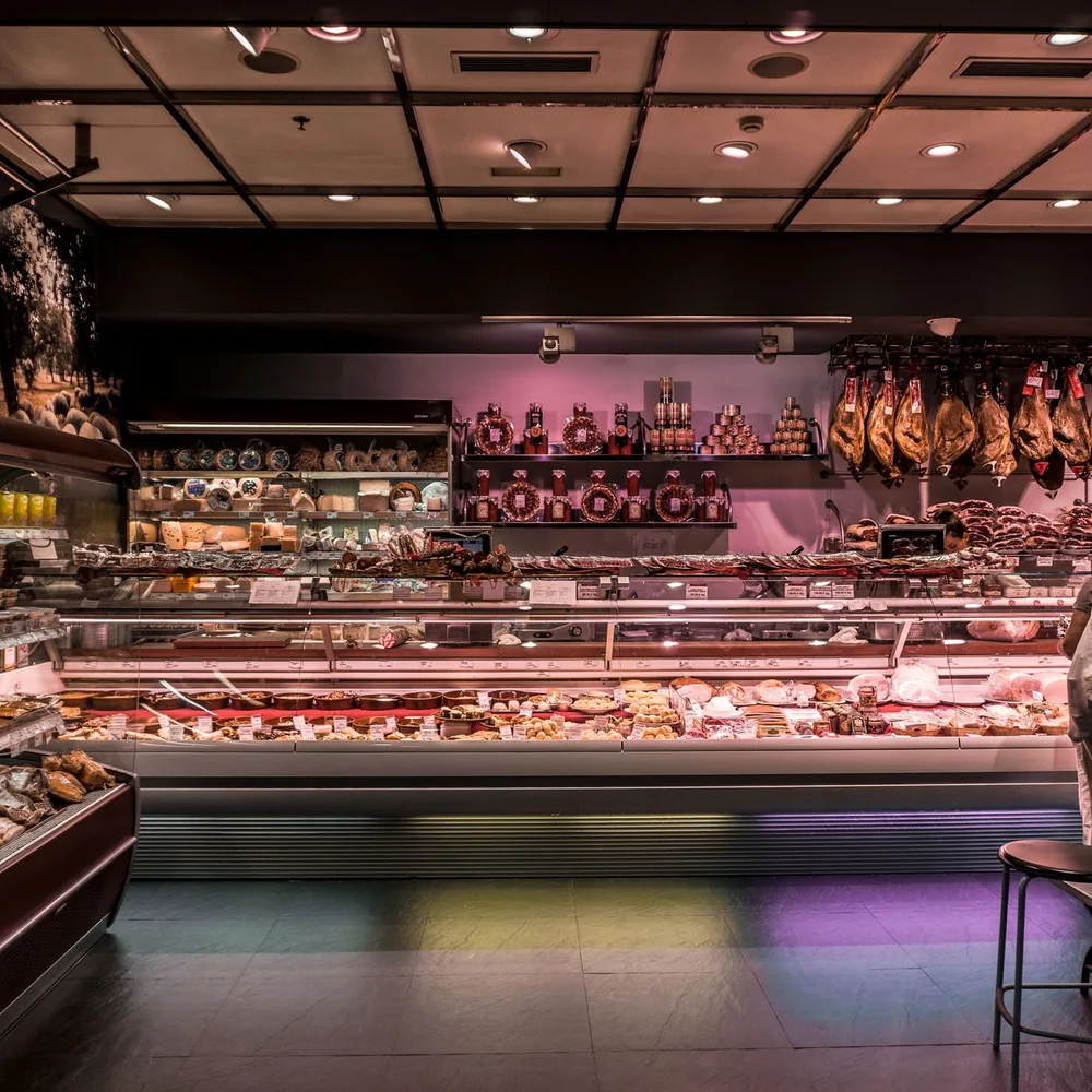 The interior of a deli with a long display counter, hanging cured meats, and shelves of food.
