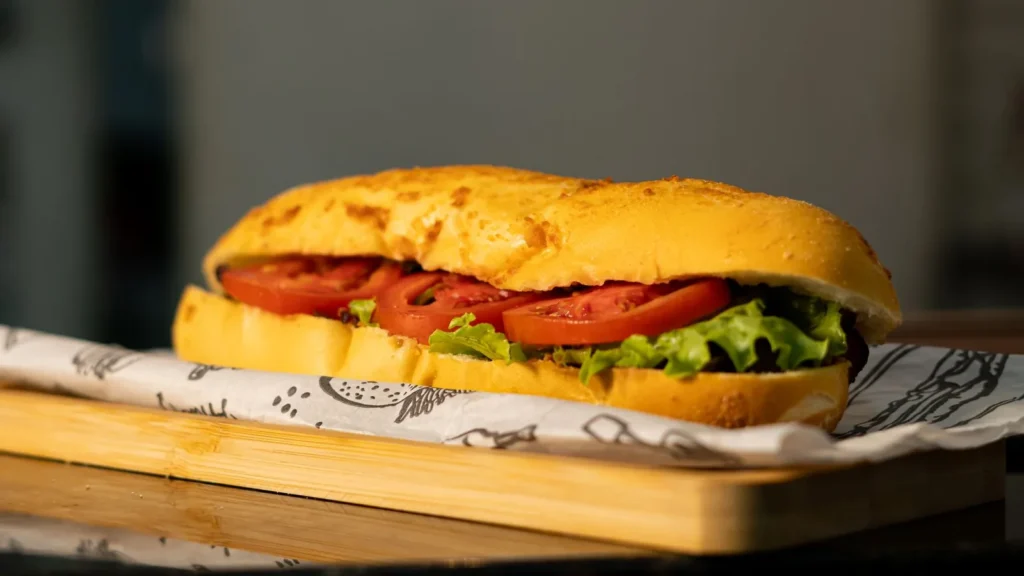 A long sandwich with tomato slices and lettuce on printed paper on a wooden board.
