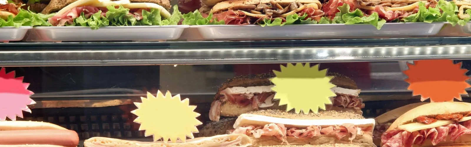 Sandwiches in a deli display case with colorful starburst signs.