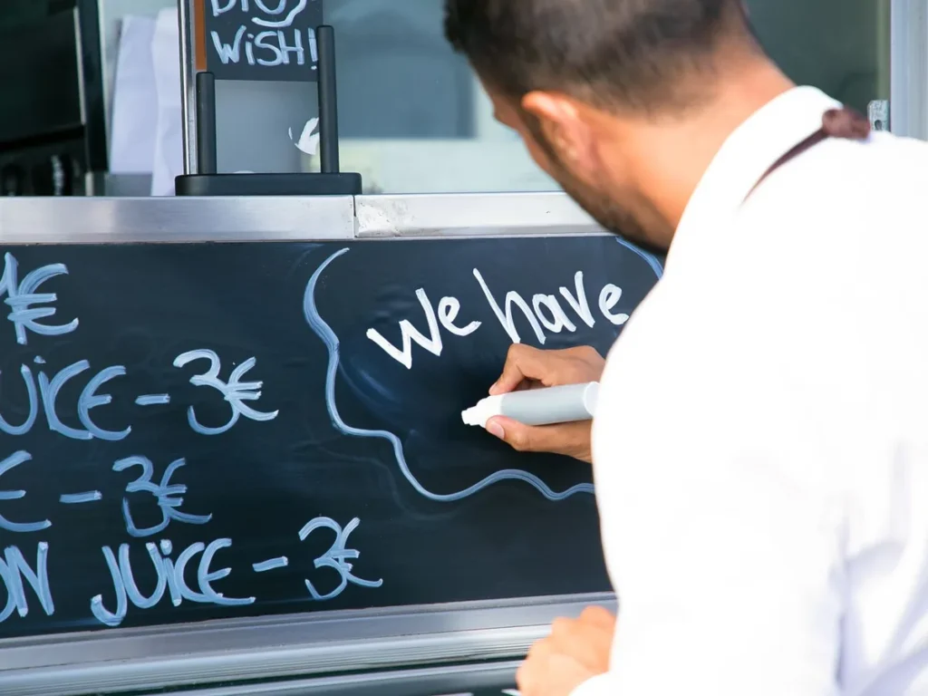 A worker writing on a chalkboard menu board listing juice prices.