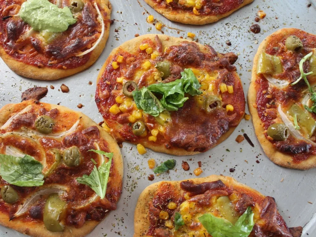 Small baked flatbreads topped with sweetcorn, olives, and green leaves on a baking tray.
